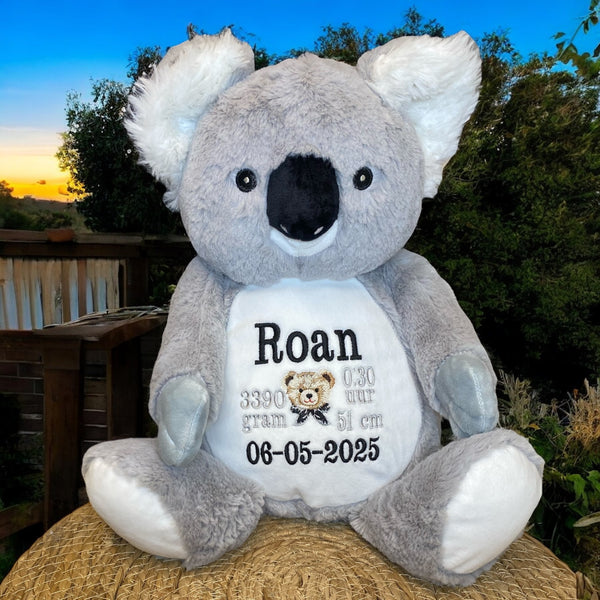 Grijze geboorteknuffel in de vorm van een koala met gepersonaliseerd borduurwerk op de buik, inclusief naam en geboortegegevens.