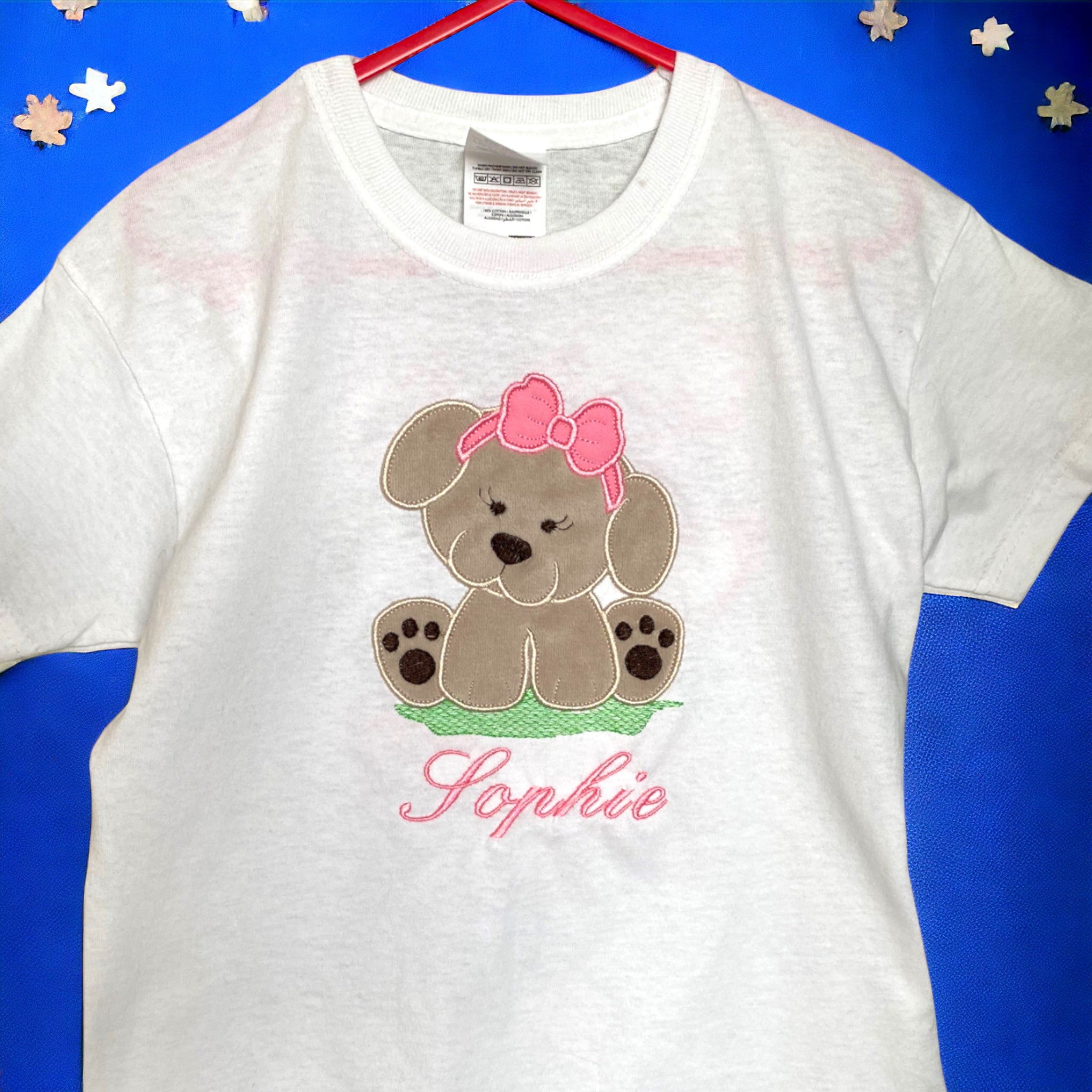 Gepersonaliseerd kinder shirt met een geborduurde applicatie van een beige hondje van Nicky Velours met een roze strik. Het hondje voelt zacht aan. Een lief, leuk, zacht kindershirt als cadeau voor de oudere zus.