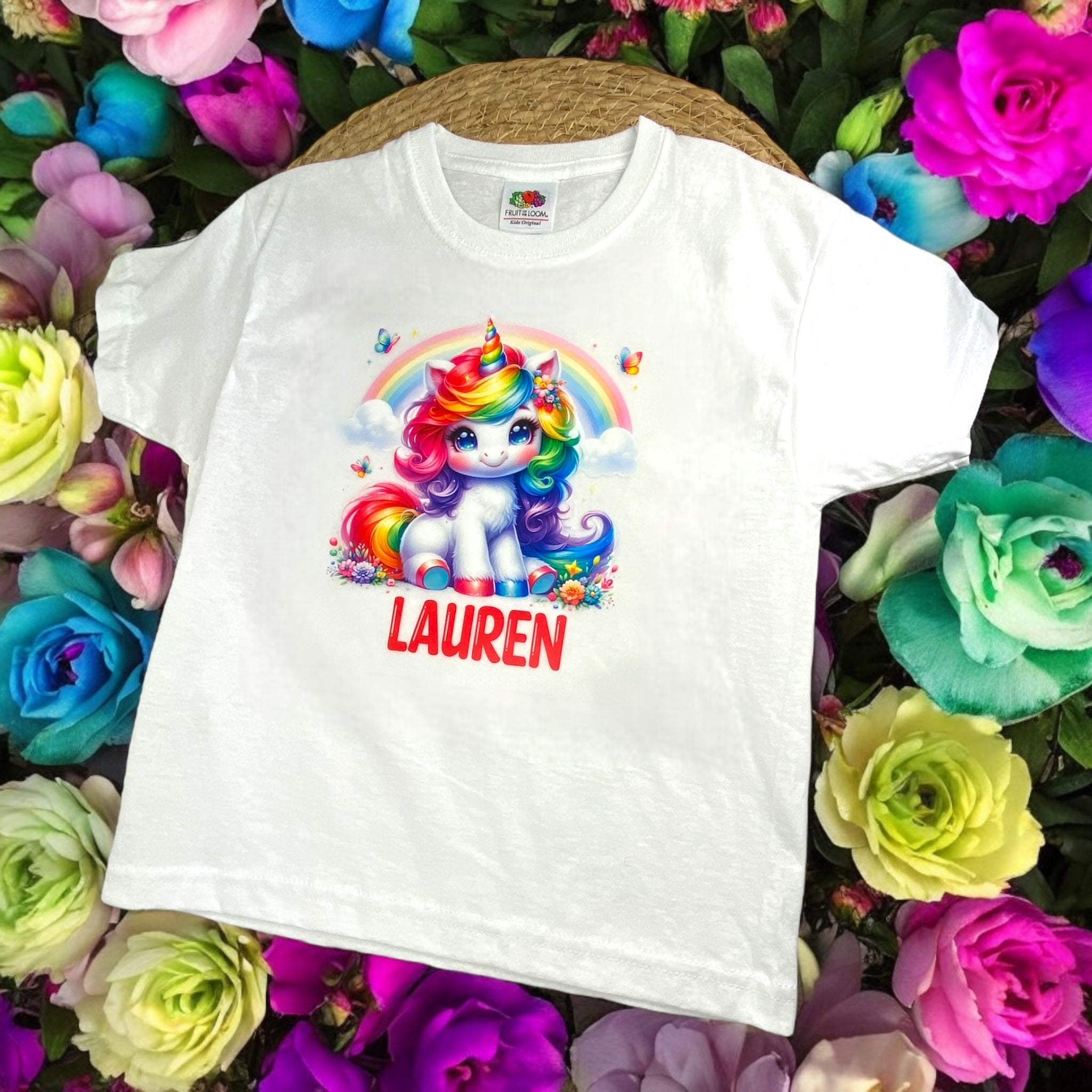 kindershirt met kleurrijke eenhoorn en regenboog, gepersonaliseerd met een naam, omringd door bloemen en vlindertjes.