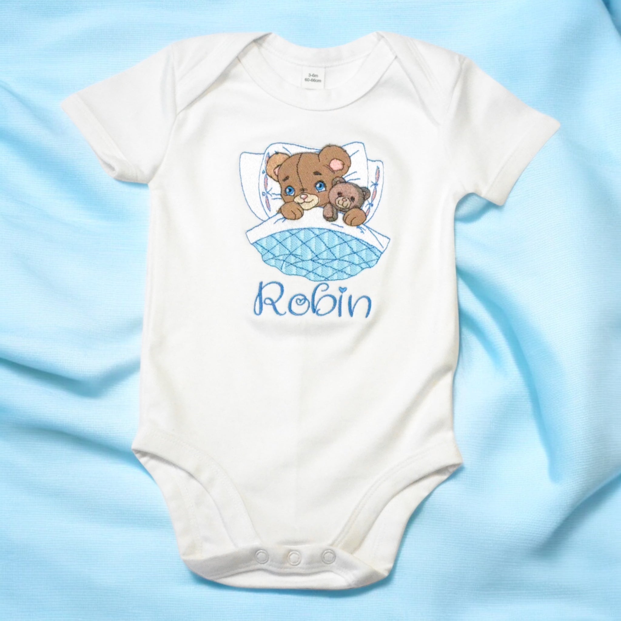 Geborduurd en gepersonaliseerd baby rompertje met een beertje die in bed ligt en een teddy beer vasthoudt. Onder het in bed liggende beertje wordt in het blauw de naam van de baby geborduurd. Echt een super leuk geboorte cadeau.