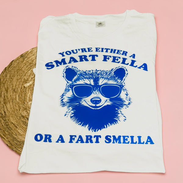 Unisex T-shirt voor volwassenen met blauwe bedrukking van een stoere wasbeer met zonnebril en de tekst "You're Either a Smart Fella or a Fart Smella", humoristisch design van SaZuDesigns.