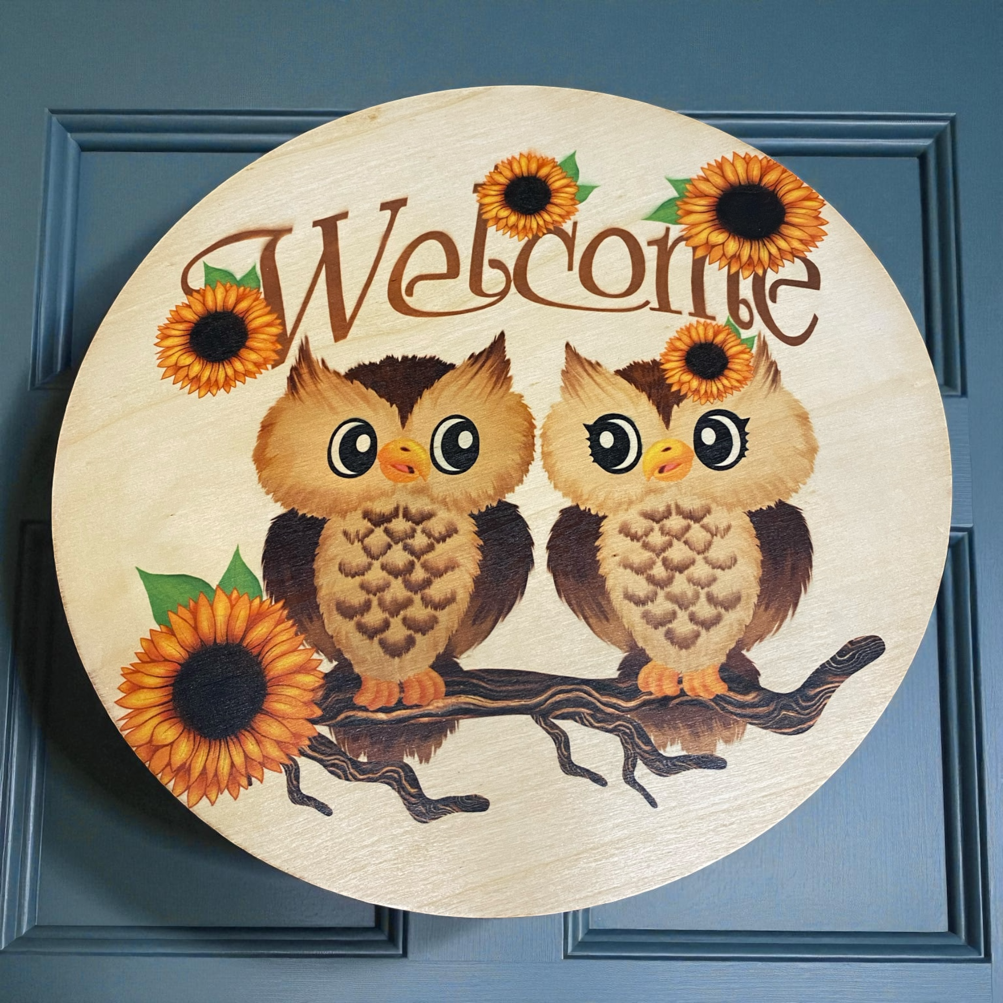 Rond houten welkomstbord met twee schattige getekende uiltjes op een tak, omringd door felgekleurde zonnebloemen, en het woord “Welcome” bovenaan in sierlijk lettertype.