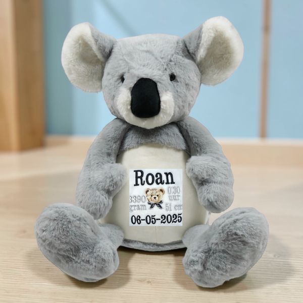 Grijze geboorteknuffel in de vorm van een koala met gepersonaliseerd borduurwerk op de buik, inclusief naam en geboortegegevens.