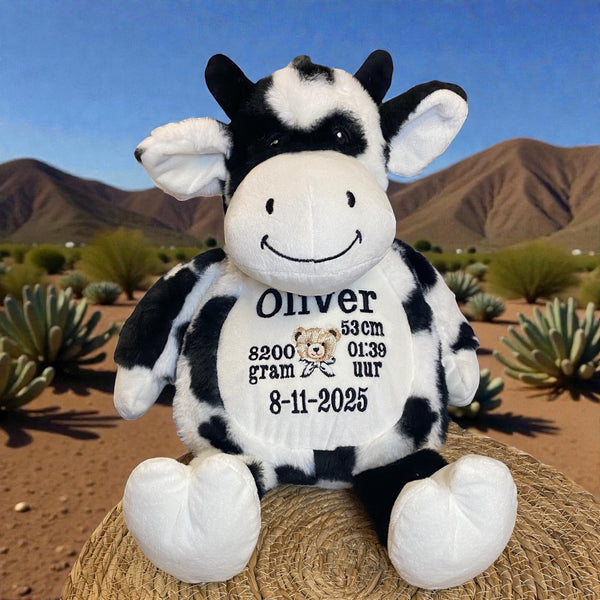 Zwart-witte geboorteknuffel in de vorm van een koe, zittend met geborduurde geboortedetails op de buik zoals naam, datum en gewicht – uniek kraamcadeau.