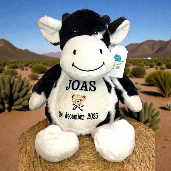 Zwart-witte geboorteknuffel in de vorm van een koe, zittend met geborduurde geboortedetails op de buik zoals naam, datum en gewicht – uniek kraamcadeau.