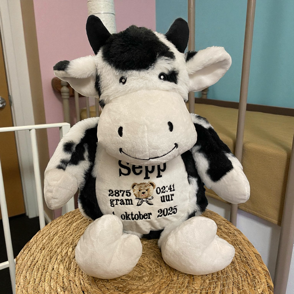 Zwart-witte geboorteknuffel in de vorm van een koe, zittend met geborduurde geboortedetails op de buik zoals naam, datum en gewicht – uniek kraamcadeau.
