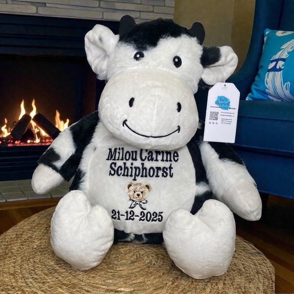 Zwart-witte geboorteknuffel in de vorm van een koe, zittend met geborduurde geboortedetails op de buik zoals naam, datum en gewicht – uniek kraamcadeau.